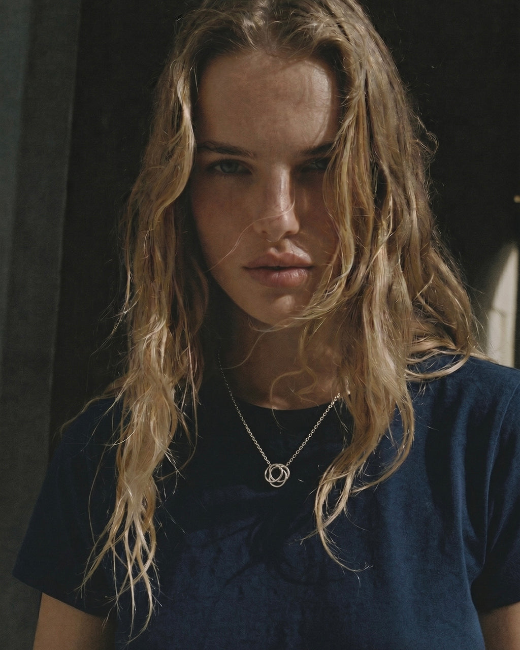 Person with long blonde hair wearing a dark blue shirt and a necklace.